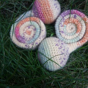 Op de afbeelding: Vier handgemaakte gehaakte slakken op gras. De slakken hebben spiraalvormige schelpen in pasteltinten roze, oranje, paars en blauw. De slakkenlichamen zijn een gespikkelde gebroken witte kleur.