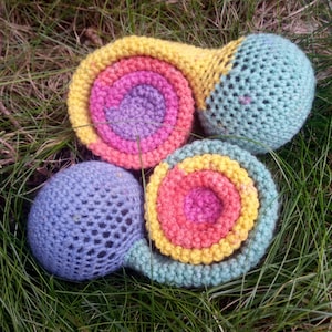 Op de afbeelding: Twee handgemaakte gehaakte slakken op gras. Eén slak heeft een spiraalvormig huisje in geel, roze en paars, met een lichtblauw lichaam. De andere heeft een huisje in geel, roze en oranje, en een paars lichaam.