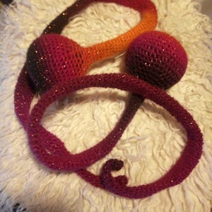 Op de afbeelding: Gehaakt kunstwerk in tinten rood, oranje en bruin, met een glitterafwerking. Het stuk heeft een grote bol en twee langwerpige, gebogen elementen. De items zijn gerangschikt op een witte, donzige ondergrond.