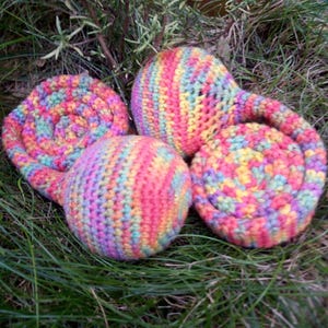 Op de afbeelding: Vier handgemaakte haakartikelen in regenboogkleuren op groen gras. Er zijn twee spiraalvormige onderzetters en twee ronde ballen. Het garen is een mix van roze, geel, blauw en groen. De items lijken handgemaakt.