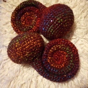 Op de afbeelding: Vier gehaakte items in tinten rood, oranje en bruin met iriserende pailletten. Twee zijn spiraalvormig en twee zijn afgerond. De items zijn gerangschikt op een witte, donzige ondergrond.