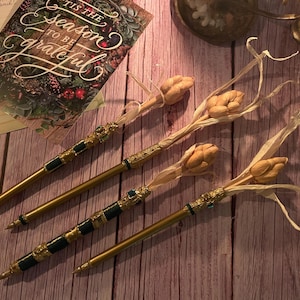 May include: Four decorative pens with gold and green accents, topped with dried flower buds and delicate leaves. The pens are arranged on a wooden surface near a greeting card that reads "'Tis the season to be grateful."