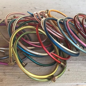 May include: A collection of colorful metal nail bracelets in various shades, including gold, silver, red, blue, and green. The bracelets are arranged in a pile, showcasing their unique nail-head design. The image is taken on a light-colored wooden surface.