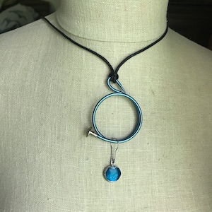 May include: A unique necklace featuring a circular blue pendant with a silver-colored nail accent. The pendant hangs from a black cord. A small, round, blue and silver charm dangles below the circle.