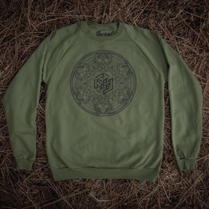 Op de afbeelding: Een groen sweatshirt met lange mouwen, een geometrisch ontwerp en het woord "Sacred" gedrukt op de hals.
