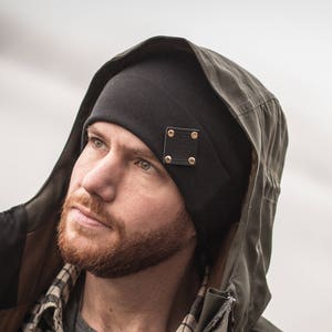 May include: A man wearing a black beanie with a leather patch and a green hooded jacket. The beanie has a gold accent on the patch.