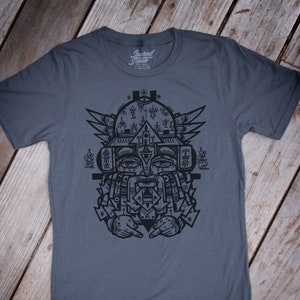May include: A dark gray t-shirt with a black graphic print of a stylized face with geometric patterns and the text "Sacred State".