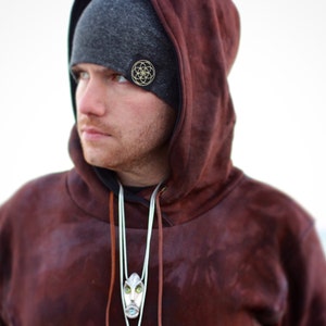 Puede incluir: Un hombre con una sudadera con capucha marr&oacute;n, una boina gris y un collar con un colgante plateado. La boina tiene un dise&ntilde;o geom&eacute;trico dorado.