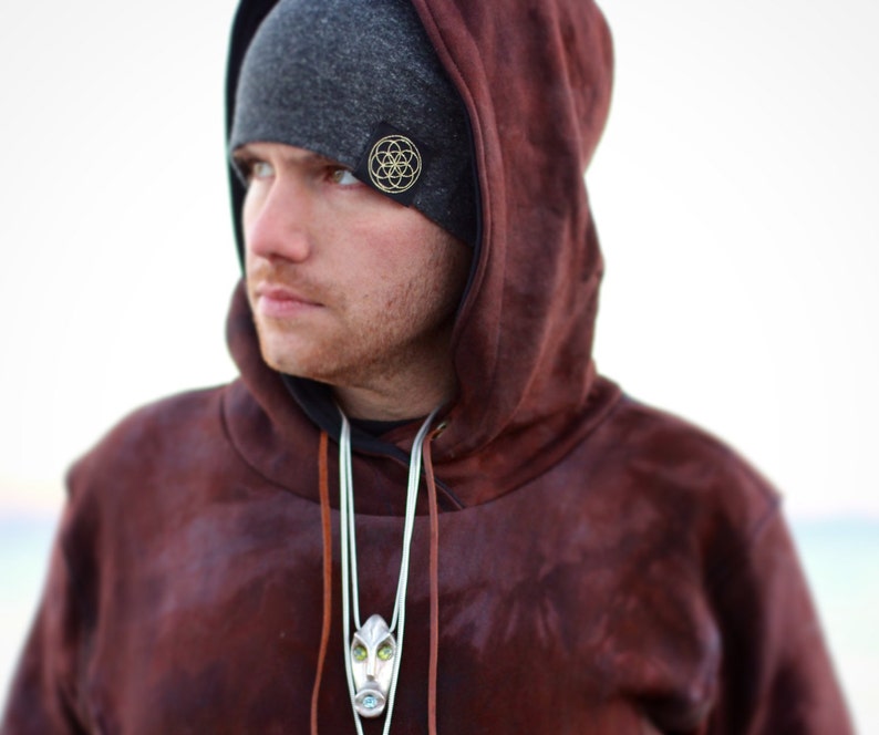 Puede incluir: Un hombre con una sudadera con capucha marr&oacute;n, una boina gris y un collar con un colgante plateado. La boina tiene un dise&ntilde;o geom&eacute;trico dorado.