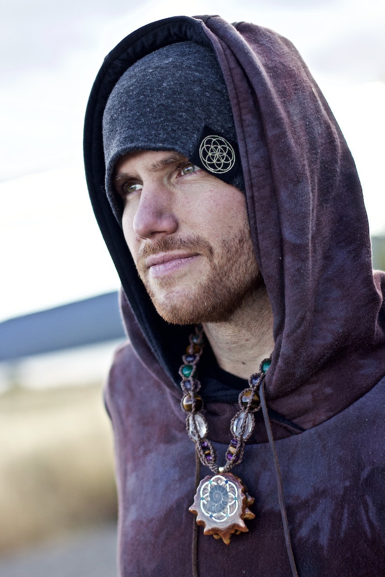 Puede incluir: Una persona lleva una boina gris oscuro con un dise&ntilde;o geom&eacute;trico dorado y una sudadera con capucha de color morado oscuro. La persona lleva un collar de cuentas con un colgante de madera con un dise&ntilde;o geom&eacute;trico similar.