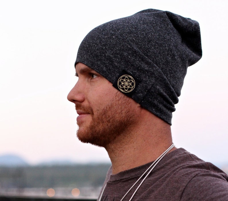 Puede incluir: Un gorro de lana gris oscuro con un peque&ntilde;o parche negro que presenta un dise&ntilde;o geom&eacute;trico dorado.