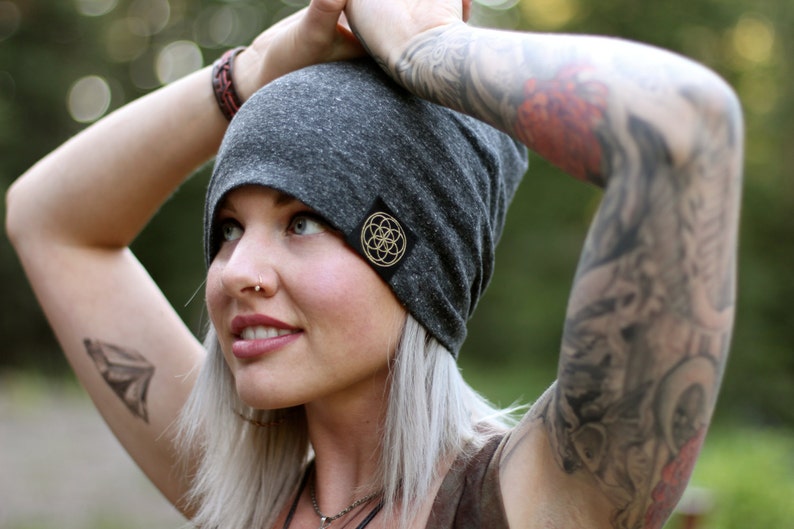 Puede incluir: Un gorro gris con un peque&ntilde;o logotipo circular dorado que presenta un dise&ntilde;o geom&eacute;trico. El gorro est&aacute; hecho de un tejido suave y el&aacute;stico y tiene un ajuste holgado.