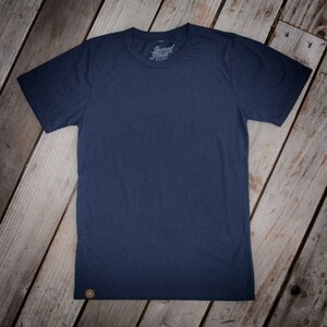May include: A navy blue t-shirt with a small, gold logo on the bottom left corner. The shirt has a crew neck and short sleeves. The text "Sacred State" is printed on the front of the shirt.