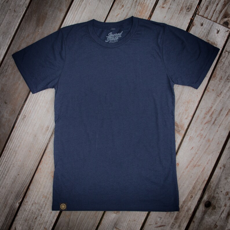 May include: A navy blue t-shirt with a small, gold logo on the bottom left corner. The shirt has a crew neck and short sleeves. The text "Sacred State" is printed on the front of the shirt.