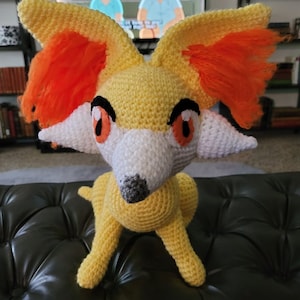 May include: A crocheted plush toy of a fox with orange fur, yellow body, and white face. The toy has black eyes and a gray nose.