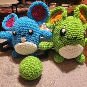 May include: Two crocheted stuffed toys, one blue with pink ears and one green with orange ears. Both have white faces with black eyes and a white belly.