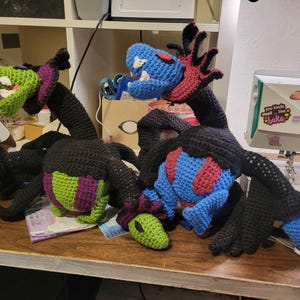 May include: Three crocheted plush toys in the shape of dragons. The dragons are black, green, and blue with red accents. The dragons are sitting on a wooden table.