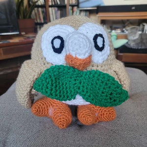 May include: A crocheted stuffed owl toy with brown, white, and green yarn. The owl is holding a green leaf in its beak.