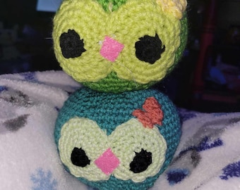 May include: Two crocheted owl plushies, one green with a yellow flower on top and one blue with an orange flower on top. Both owls have black eyes and pink noses.