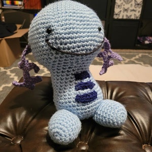 May include: A crocheted blue and purple plush toy with a smiling face and three purple stripes on its body.