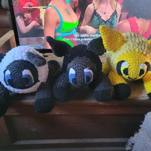 May include: Three crocheted stuffed animals: a white sheep with black accents, a black sheep with white accents, and a yellow sheep with black accents. All three have blue eyes.