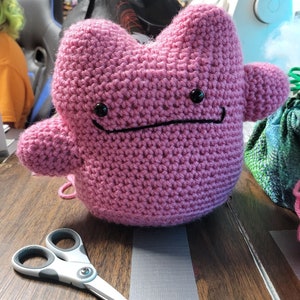 May include: A pink crocheted plushie with black eyes and a black mouth. The plushie is sitting on a wooden table with a pair of scissors next to it.