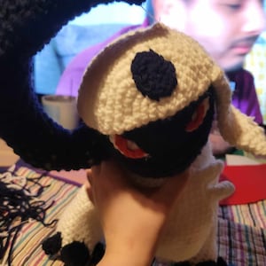 Peut inclure: Un jouet en peluche crocheté ressemblant à un personnage de Pokemon. Le jouet est blanc avec des accents noirs et une queue en forme de croissant de lune. Le jouet a des yeux rouges et un nez noir.