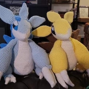 May include: Two crocheted stuffed toys, one blue and white and one yellow. The blue and white toy is sitting on a brown surface with a ball of yarn next to it. The yellow toy is sitting on the brown surface with its legs crossed.