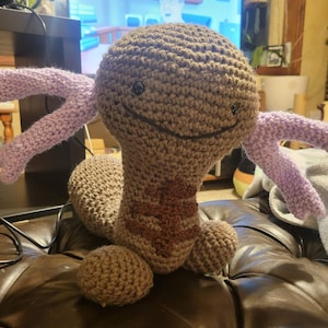 May include: A handmade crochet plush toy in shades of brown and purple. The toy has a rounded head, a long body, and two small feet. It features two purple tentacles and a smiling face with black eyes and a stitched mouth.