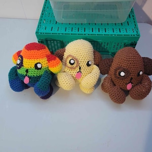 Puede incluir: Tres juguetes de cachorros de ganchillo. Uno es de color arcoíris, otro es amarillo con orejas marrones y el último es marrón. Cada uno tiene ojos blancos, una lengua rosa y una expresión sonriente. Están sobre una superficie blanca.