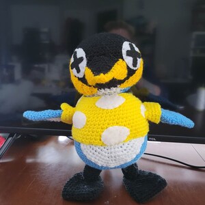 May include: A crocheted plush toy penguin with black and white eyes, a yellow beak, and a yellow and white polka dot shirt. The penguin is standing on a wooden surface.