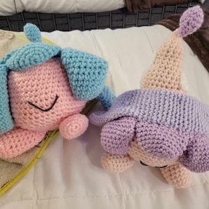 Puede incluir: Dos animales de peluche tejidos a crochet, uno rosa con orejas azules y otro morado con un sombrero rosa. Ambos están acostados sobre una superficie blanca.