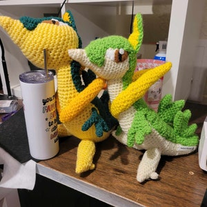 Peut inclure: Deux jouets en peluche crochetés, l'un jaune et vert, l'autre vert et blanc, sont posés ensemble sur une surface en bois. Le jouet jaune et vert a un long museau et une crête verte. Le jouet vert et blanc a un long museau et une collerette verte autour du cou. Les deux jouets se tiennent l'un l'autre.