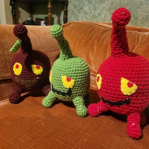 May include: Three handmade crochet monster plushies in burgundy, green, and red, each with yellow eyes and a black stitched mouth. The burgundy monster has a green leaf on top. They are sitting on an orange couch.