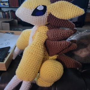 May include: A crocheted plush toy in the shape of a yellow and brown creature with white spikes. The toy has a blue eye and is sitting on a brown sofa.