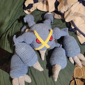 Puede incluir: Peluche de Metagross tejido a ganchillo en tonos azules, grises, amarillos y rojos. El peluche presenta un diseño detallado con múltiples extremidades y una distintiva cruz amarilla en la cabeza. Hecho a mano.