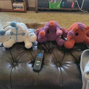 May include: Three handmade crochet octopus plush toys in white, purple, and orange. The white octopus has blue accents, while the purple and orange ones have contrasting color details. A black remote control is in the foreground.