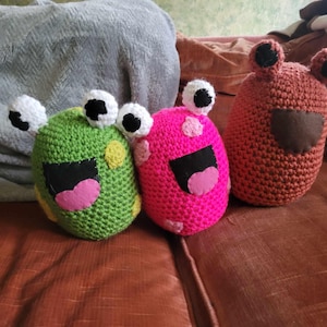 May include: Three handmade crochet monster toys in green, pink, and brown. Each has large white eyes with black pupils, a black mouth, and a pink tongue. The green and pink monsters have yellow spots.