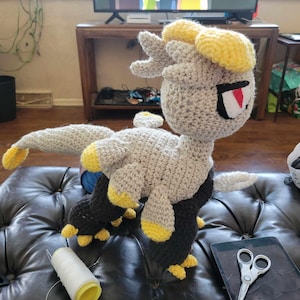 Pode incluir: Um dragão de crochê cinza e preto com detalhes amarelos. O dragão tem um olho vermelho e está sentado em um sofá de couro preto.