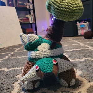 May include: A crocheted plush toy of a brown and green turtle with a green tree on its back. The turtle has red eyes and a white belly.