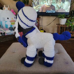 May include: A white crocheted plush toy with blue accents. The toy is wearing a blue and white striped sailor hat. The toy has a long tail and is standing on a light gray surface.