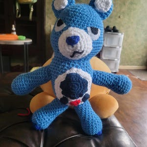 May include: A blue crocheted teddy bear with white accents and a red heart on its chest. The bear has a grumpy expression and is sitting on a brown leather sofa.