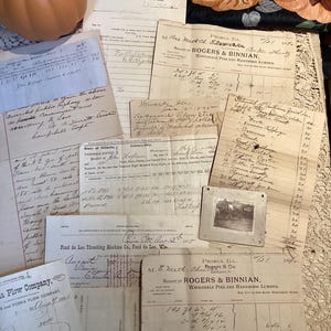 May include: A collection of antique paper documents, including handwritten receipts, invoices from Rogers & Binnian, and a Plow Company form, layered with a small square black and white photograph and a decorative orange pumpkin.