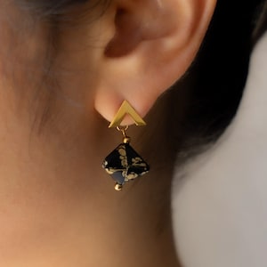 May include: Gold earrings with a black and gold geometric design. The earrings feature a small triangle at the top and a larger square-shaped pendant with a black and gold pattern.