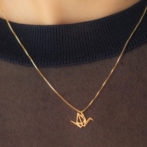 May include: A delicate gold necklace featuring a geometric origami crane pendant. The pendant is suspended from a fine chain, creating a minimalist and elegant design. The necklace is shown against a dark background.