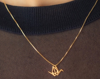 Peut inclure: Un collier doré délicat avec un pendentif grue origami géométrique. Le pendentif est suspendu à une fine chaîne, créant un design minimaliste et élégant. Le collier est présenté sur un fond sombre.