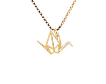 Peut inclure: Un collier doré avec un pendentif grue origami géométrique. Le pendentif est suspendu à une chaîne délicate avec un motif à maillons carrés. Le collier est présenté sur fond blanc.
