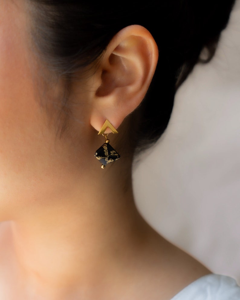 May include: Gold earrings with a black and gold geometric design. The earrings feature a small triangle at the top and a larger square-shaped pendant with a black and gold pattern.