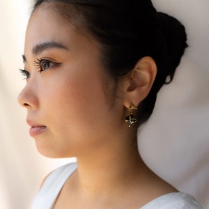 May include: A woman with dark hair is wearing gold earrings with a black and gold geometric design.