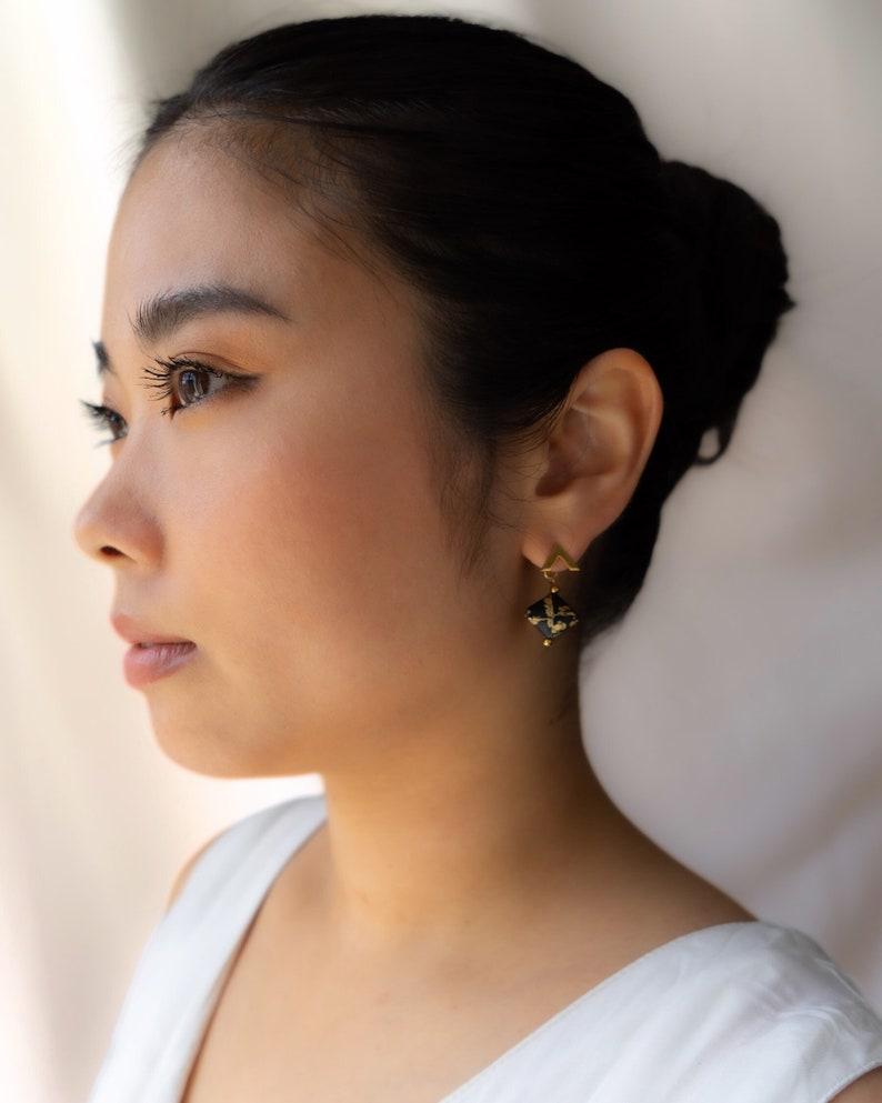 May include: A woman with dark hair is wearing gold earrings with a black and gold geometric design.